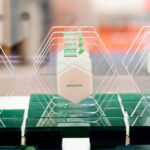 Três troféus hexagonais de acrílico transparente com o logotipo BRDE e inscrição "DUALPIXEL" estão alinhados sobre uma superfície verde.
