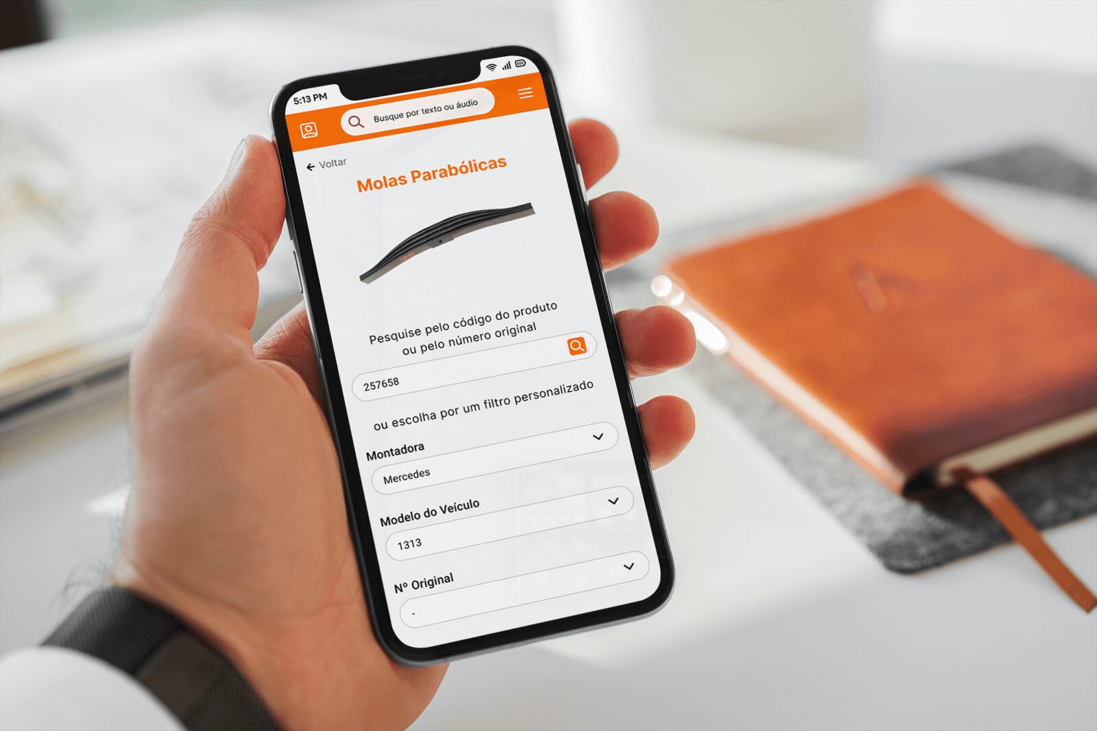 Mão segurando um smartphone que exibe a tela de busca do aplicativo da AESA na seção ‘Molas Parabólicas’. No topo, há imagem de uma mola e, abaixo, campo para pesquisar pelo código do produto ou número original, com exemplo preenchido ‘257658’. Abaixo, há opções de filtro personalizável como ‘Montadora’ (Mercedes), ‘Modelo do Veículo’ (1313) e ‘Nº Original’. Ao fundo, sobre a mesa desfocada, há um caderno de couro marrom e outros objetos de escritório.