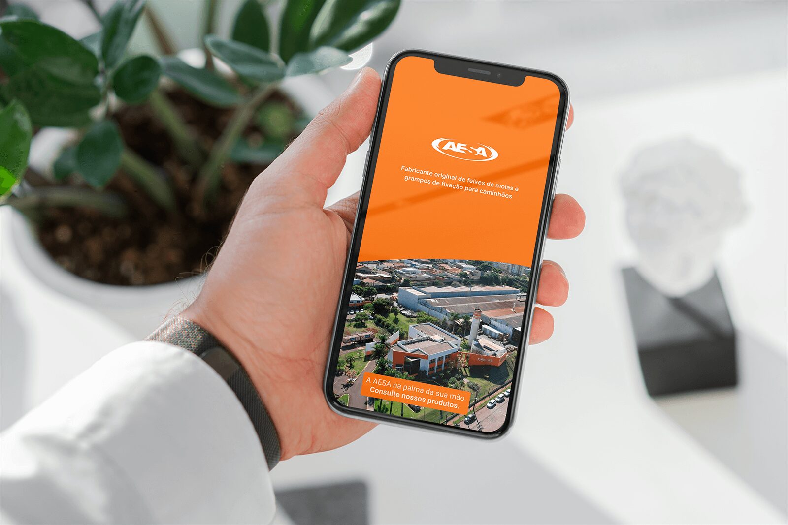Mão segurando um smartphone que exibe a tela inicial do aplicativo da AESA. A parte superior da tela é laranja, com o logotipo da empresa e o texto ‘Fabricante original de feixes de molas e grampos de fixação para caminhões’. A parte inferior mostra uma foto aérea do complexo industrial da AESA, acompanhada da frase ‘A AESA na palma da sua mão. Consulte nossos produtos.’ em destaque laranja. Ao fundo, desfocado, há um vaso de planta e uma escultura branca.